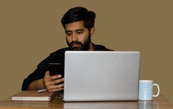 Молодий індійський чоловік дивиться на телефон під час роботи на ноутбуці. Вибірковий фокус на обличчі людини
. - Фото, зображення