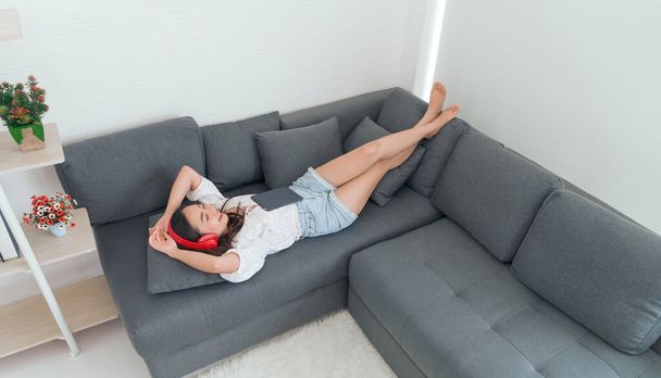 Jovem mulher asiática usar fones de ouvido vermelhos relaxar e ouvir a música ao usar tablet deitado no sofá na sala de estar, menina gosta de som de áudio, relaxante - Foto, Imagem