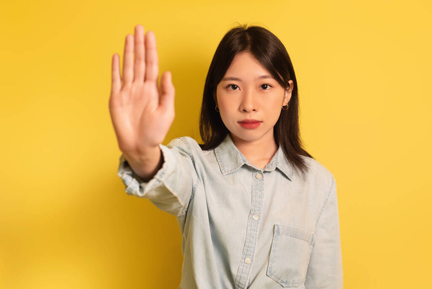 Young Asian lady gesturing STOP, showing palm at camera on yellow studio background - Fotografie, Obrázek
