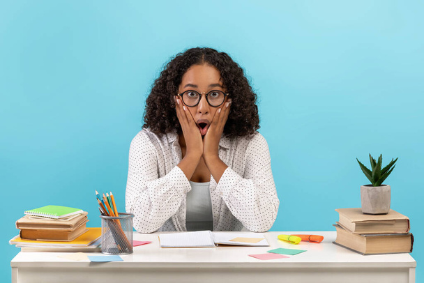 Shocked young black woman in eyeglasses sitting at table with study materials on blue studio background - 写真・画像