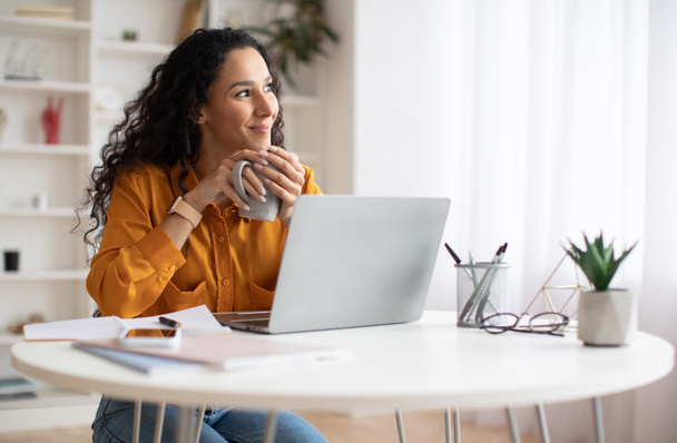 Dreamy Businesswoman Using Laptop And Holding Coffee Cup Indoors - Zdjęcie, obraz