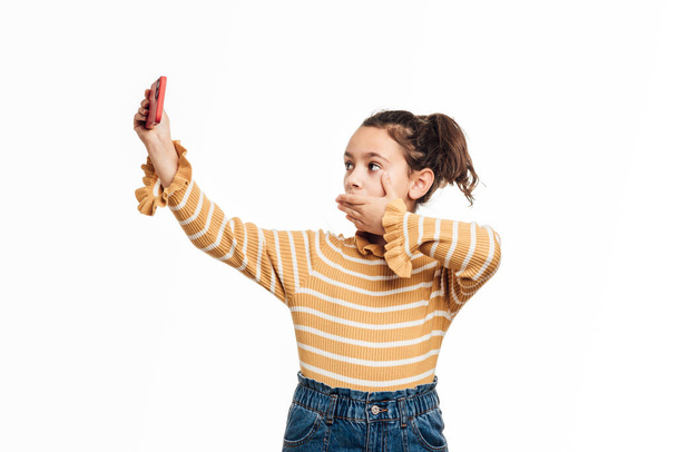 Girl covering her mouth with hand while taking a selfie with a mobile phone over an isolated background. - Foto, Imagen
