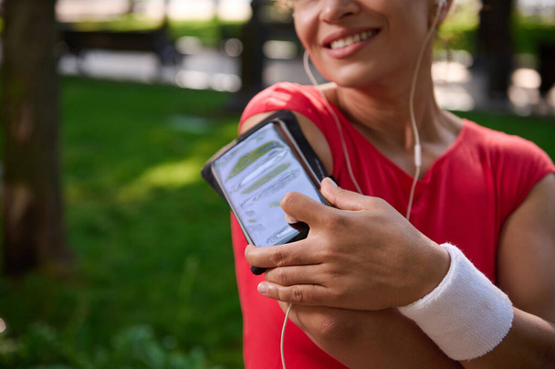 Zachte focus op een smartphone houder op de arm van wazig charmante glimlachende vrouwelijke atleet, het zetten van haar vinger op de telefoon en het aanpassen van mobiele app en afspeellijst tijdens de ochtend joggen. Sport en gadgets - Foto, afbeelding