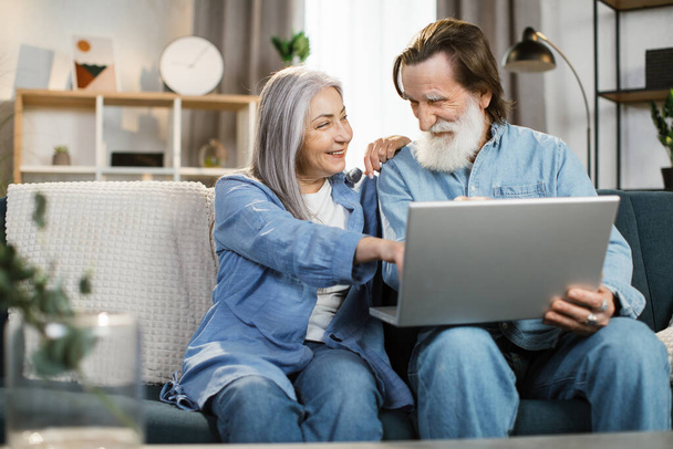 Happy mature family in casual outfit sitting together on couch and using wireless laptop. - 写真・画像