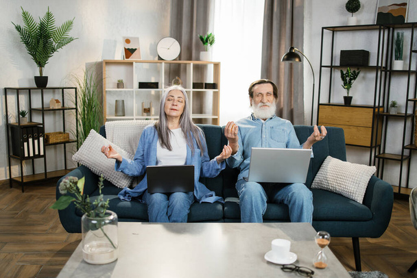 Mature woman and her bearded husband sitting on couch with laptops on knees and meditating. - Photo, image
