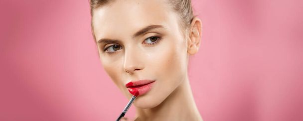 Beauty Concept - vrouw rode lippenstift met roze studio achtergrond toe te passen. Mooi meisje maakt make-up. - Foto, afbeelding