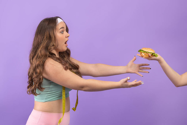 hambriento divertido joven europeo más tamaño mujer en ropa deportiva alcanza para hamburguesa en la mano - Foto, Imagen