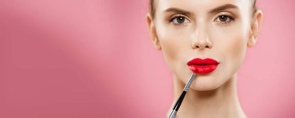 Beauty Concept - Woman applying red lipstick with pink studio background. Beautiful girl makes makeup. - Фото, изображение