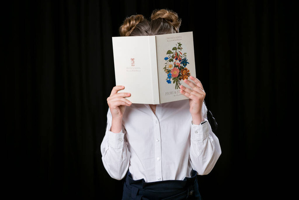 Ένα κορίτσι με λευκό πουκάμισο κάθεται σε μια καρέκλα. Το κορίτσι κλείνει τα μάτια της με ένα βιβλίο. Καλή διάθεση.. - Φωτογραφία, εικόνα