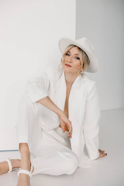 Confident young blond woman smiling, looking at camera isolated on white background. Studio portrait of successful friendly female in white suit and hat, posing over white wall. - Foto, immagini