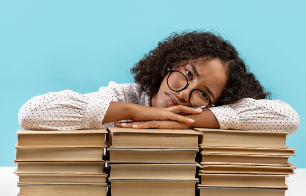 Sleepy young black female student leaning on big stacks of books, tired of getting ready for exam, feeling bored - Фото, изображение