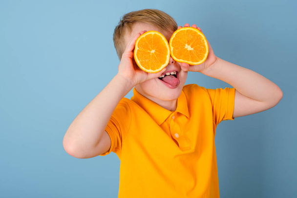 Pozytywne emocje. Zaskoczony chłopak z pomarańczą na oczach. Wesołe dziecko trzymające dłońmi dwie połówki pomarańczy. Odizolowany na niebieskim tle. Dzieciak w żółtej koszulce. Wysokiej jakości zdjęcie - Zdjęcie, obraz