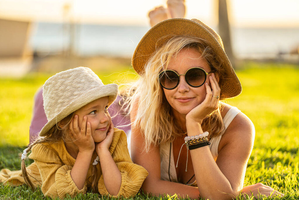 Happy mother and her little daughter having fun together on the green grass. Lovely kid lying and relaxing with her mom during summer vacation. Real people emotion. Family lifestyle. - Φωτογραφία, εικόνα