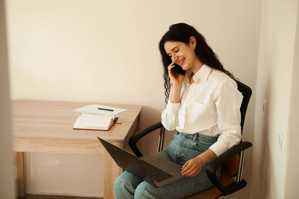 Radosna menadżerka z laptopem na kolanach rozmawiająca przez telefon, odwalająca papierkową robotę. Dziewczyna szef uśmiecha się i pracuje w biurze - Zdjęcie, obraz