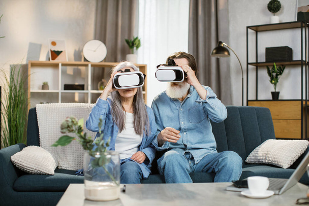 Pleasant old wife and her bearded husband in VR glasses sitting on sofa at living room at home. - Fotoğraf, Görsel