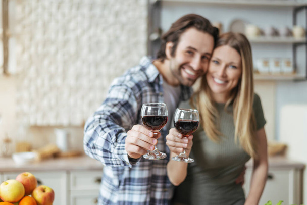 Glad millennial european couple hugs, clking glasses of wine and look at camera at scandinavian kitchen interior - Фото, изображение