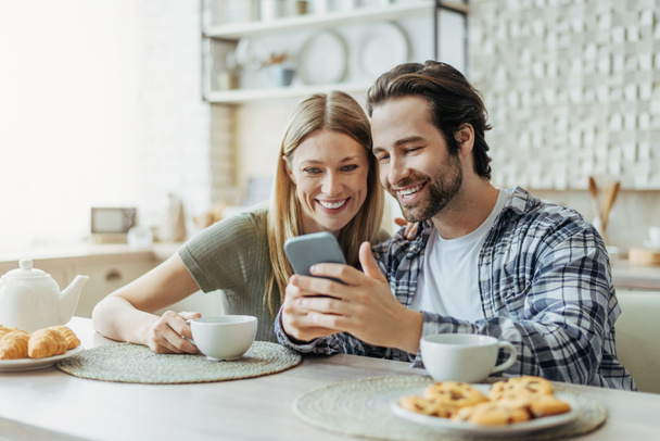 Mosolygó fiatal európai család néz okostelefon, csevegés a szociális hálózatok, igyon teát a modern konyhában - Fotó, kép