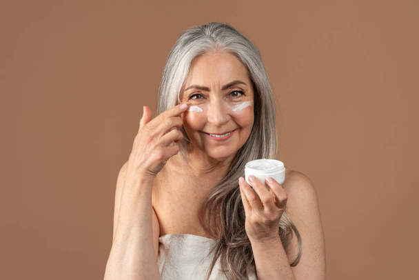 Krásná starší žena použití hydratační nebo vyživující krém pod jejíma očima na hnědém pozadí studia - Fotografie, Obrázek