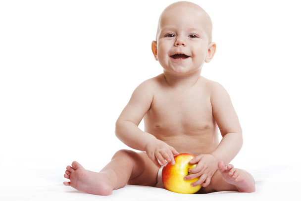 Happy child baby boy sitting in diaper and eating red yellow apple blue eyes looking at the corner isolated on a white background - Photo, Image