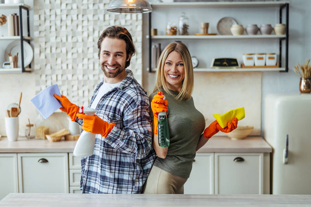 Glimlachende jonge Europese dame en man in rubberen handschoenen staan rug aan rug met schoonmaakmiddelen in handen - Foto, afbeelding