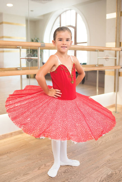 Cute portrait of a beautiful little ballerina in a performance red dress with pink tutu. She smiles because she is happy to become a professional ballerina in the future - Photo, Image