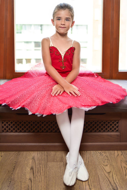 Plně dlouhý portrét krásné malé baletky v provedení červené šaty sedí u okna a sní stát se profesionální baletní tanečnice - Fotografie, Obrázek