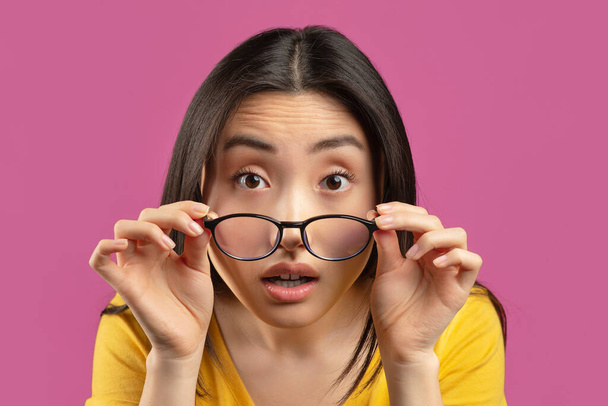 Pobre vista. Asiática dama usando gafas y mirando a la cámara por encima de las gafas, de pie sobre violeta estudio fondo - Foto, imagen