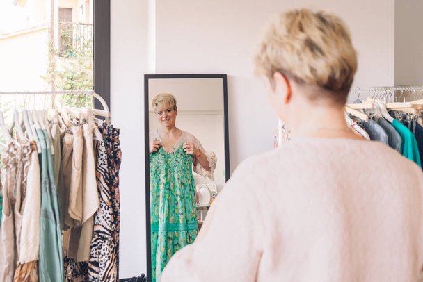 dospělá žena zkouší oblečení před zrcadlem. blondýnka nakupuje v módním obchodě. nákupní koncept. - Fotografie, Obrázek