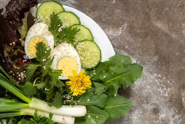 Πράσινα φρέσκα βότανα, βραστό αυγό και αγγούρι σε λευκό πιάτο σε τσιμεντένιο φόντο. Τσουκνίδα, πικραλίδα, μαρούλι, πράσινο κρεμμύδι. Μαγειρεύω καθαρό φαγητό. Θέα από ψηλά. αντίγραφο χώρου.  - Φωτογραφία, εικόνα