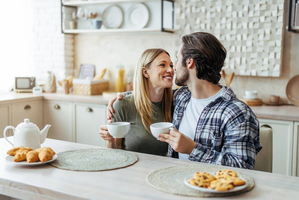 Happy young caucasian couple look at each other, drink tea, enjoy free time at weekend in modern kitchen - Фото, зображення