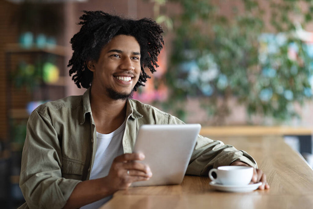 Smiling millennial black curly male blogger with tablet sitting at table, drinking coffee and look at free space - Foto, Imagem
