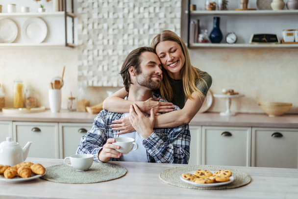 Glad young european lady hugging male with stubble drink coffee, enjoy free time in modern kitchen interior - 写真・画像