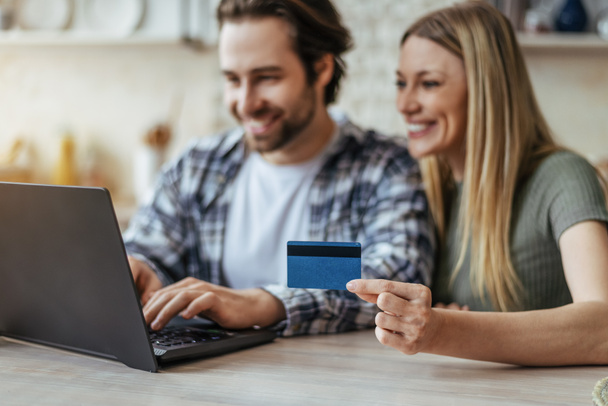 Glad young caucasian couple shopping online and show credit card in kitchen interior, blurred - Photo, image