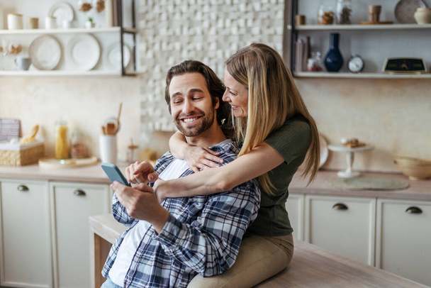Happy young european wife hugging her husband, guy shows smartphone in modern kitchen interior - Foto, Imagen