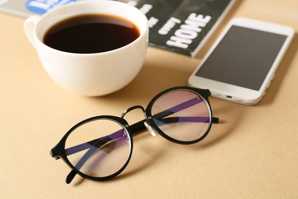 Eyeglasses, mobile phone, cup of coffee and magazine on beige background, closeup - Fotografie, Obrázek