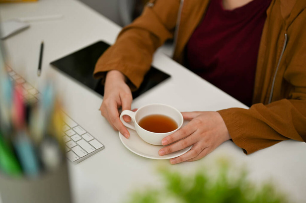 Trabajadora o diseñadora gráfica sentada en el escritorio de su oficina, tomando un té de la tarde en la oficina. - Foto, Imagen