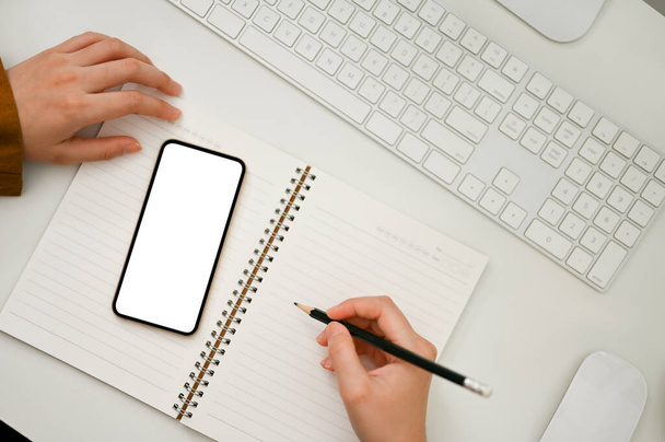 Vue de dessus, Femme travaillant dans son espace de travail, écrivant sur un carnet spirale vide et utilisant un smartphone. téléphone écran blanc maquette. - Photo, image
