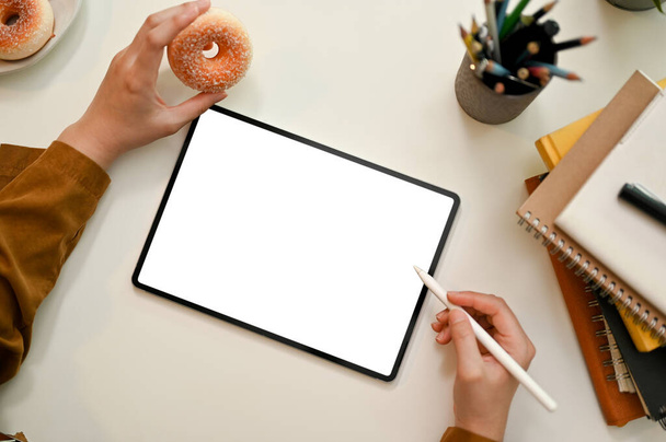 Vista superior, manos femeninas sosteniendo un pedazo de rosquilla y utilizando lápiz lápiz lápiz de dibujo en la pantalla de la tableta. Tableta mockup pantalla blanca. - Foto, imagen