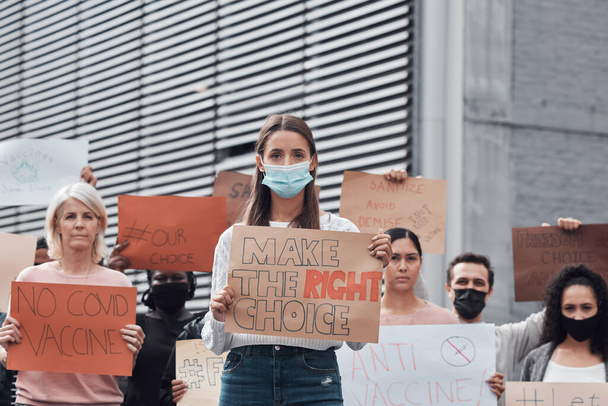 Defendiendo los derechos humanos. Retrato recortado de una atractiva joven sosteniendo un cartel protestando contra la vacuna covid 19 con otros manifestantes en el fondo. - Foto, imagen