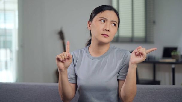 Asian woman feel confused shrugging and pointing up sitting in living room at home. - Zdjęcie, obraz