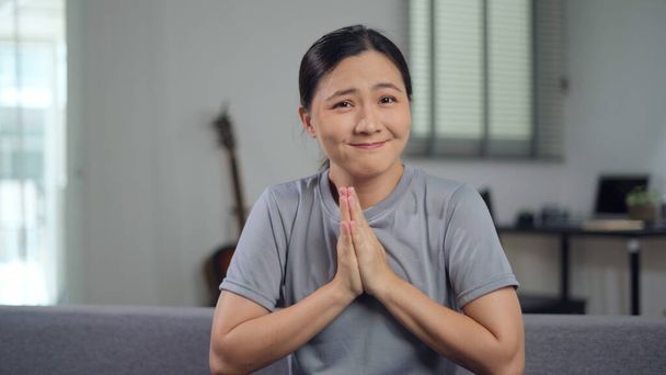 Mujer asiática cogida de la mano en oración, mirando a la cámara sentada en el sofá en la sala de estar en casa. - Foto, imagen