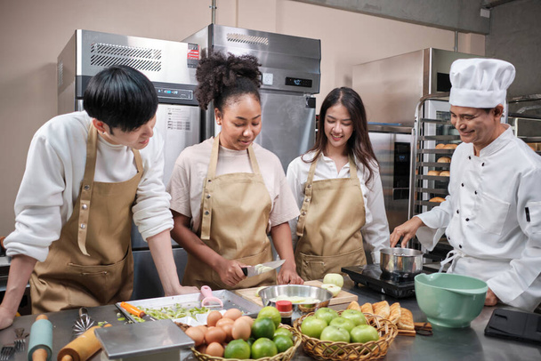 Hobby cuisine course, senior male chef in cook uniform teaches young cooking class students to peel and chop apples, ingredients for pastry foods, fruit pies in restaurant stainless steel kitchen. - Foto, Bild