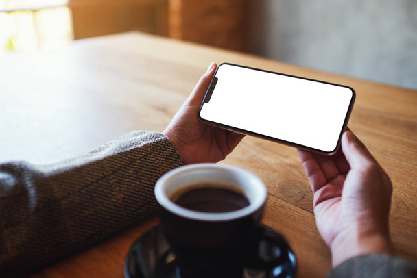 Mockup image of a woman holding black mobile phone with blank screen with coffee cup on wooden table - Fotografie, Obrázek
