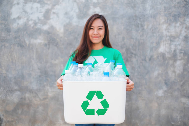 Una mujer asiática sosteniendo una papelera de reciclaje con botellas de plástico de pie frente al fondo de la pared de hormigón - Foto, Imagen