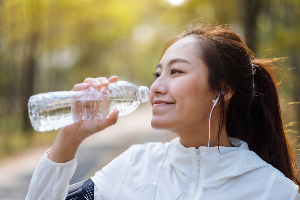 Imagen de cerca de una corredora asiática bebiendo agua de la botella después de trotar en el parque de la ciudad - Foto, Imagen