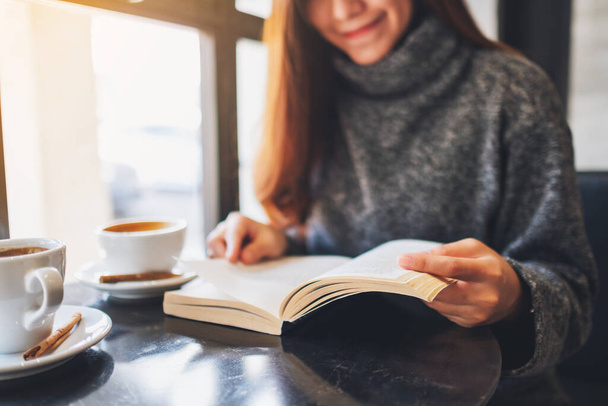 Крупним планом зображення красивої азіатки тримає і читає книгу з чашкою кави на столі
 - Фото, зображення