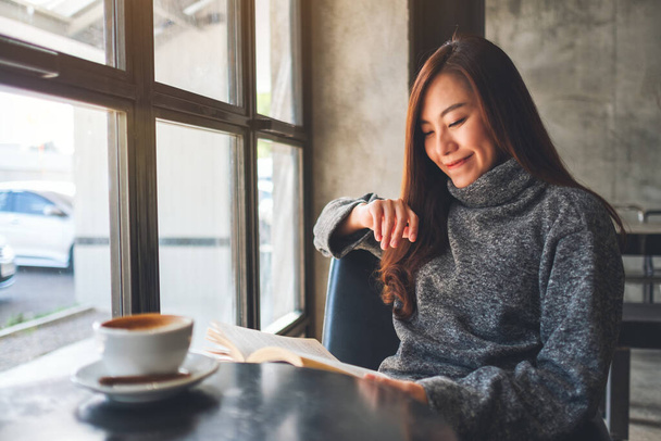 Κοντινή εικόνα μιας όμορφης Ασιάτισσας που κρατάει και διαβάζει ένα βιβλίο με ένα φλιτζάνι καφέ στο τραπέζι - Φωτογραφία, εικόνα
