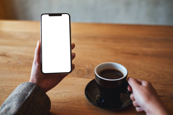 Mockup afbeelding van een vrouw met een mobiele telefoon met blanco wit scherm tijdens het drinken van koffie - Foto, afbeelding