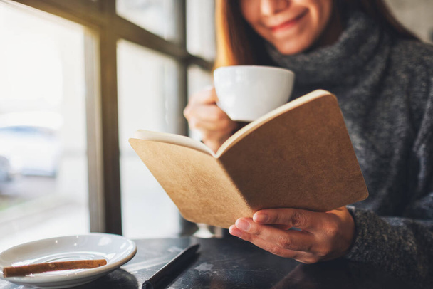 Closeup image of a beautiful woman reading a book while drinking coffee in the morning - Фото, изображение