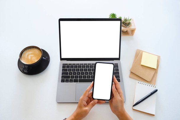 Изображение рук, держащих белый экран мобильного телефона и ноутбука на столе в офисе - Фото, изображение